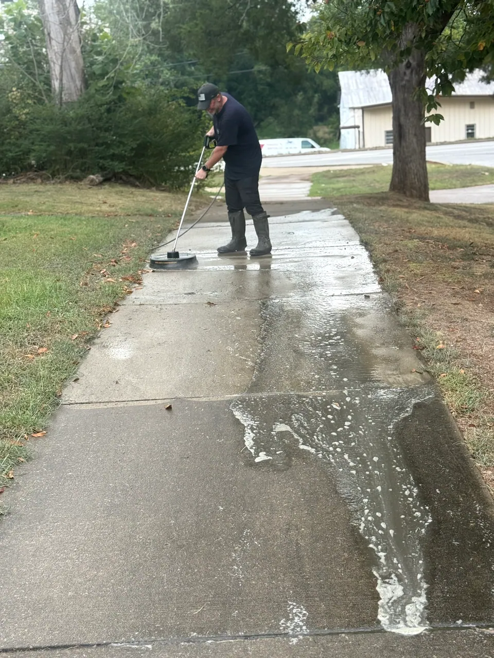 Operator moving a surface cleaner along a wet concrete sidewalk bordered by grass